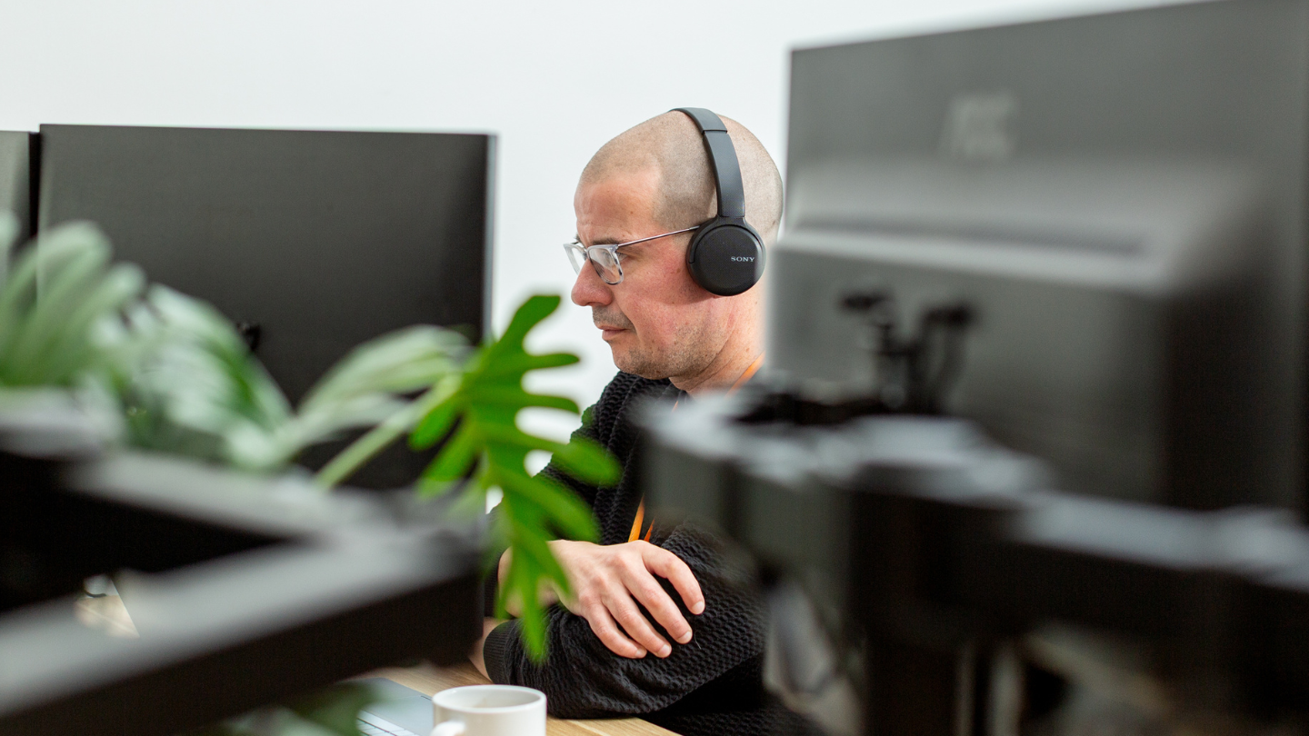 IT consultant Lee working at his computer with headphones on and plant in the foreground