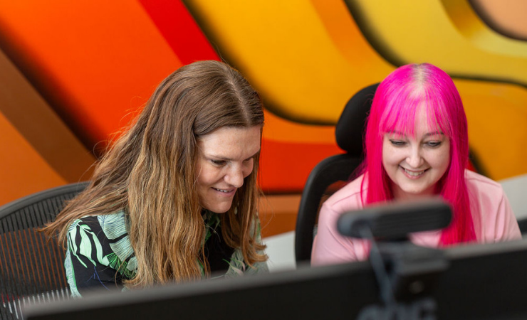 Two staff members chatting next to a computer. Background the bright Resolve waves.