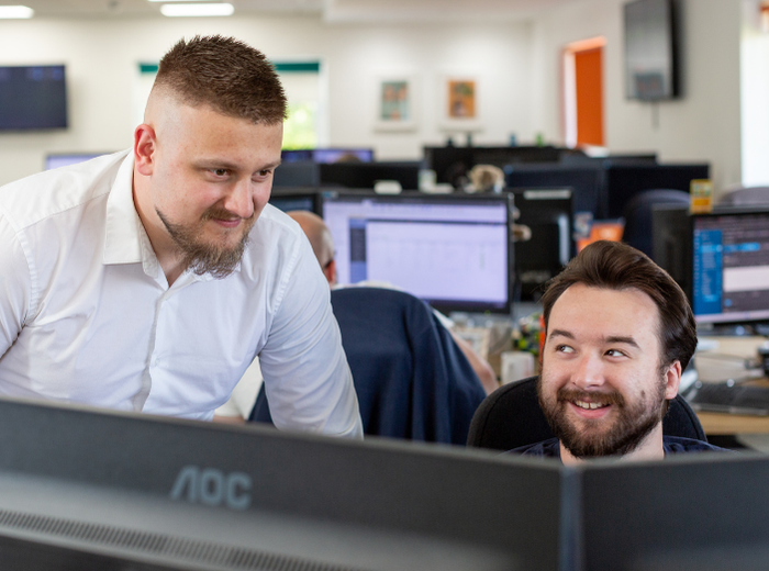 Photo of two staff members smiling at each other whilst sat behind a computer.