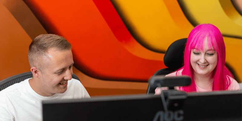 2 staff members at a computer. Man with shortlight brown hair and a women with long pink hair.